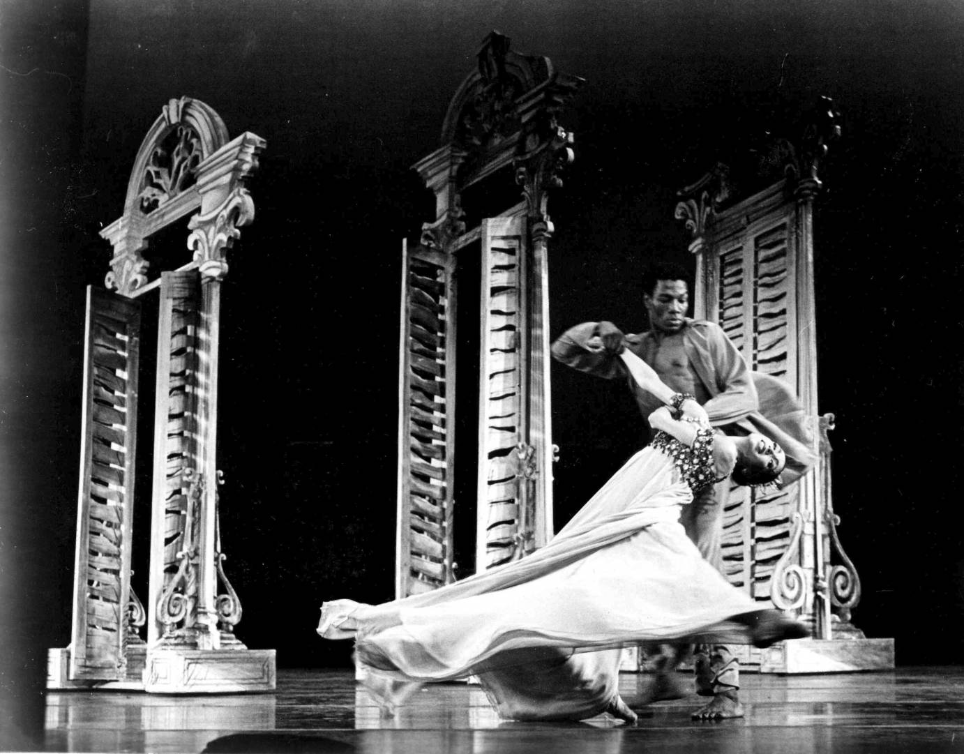 a muscular, Lowell Smith sweeps Virginia Johnson around his lower body, in the ballet. On stage they dance in front of a set of louvered windows